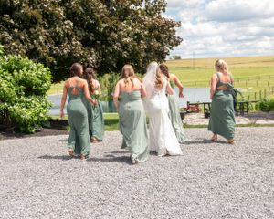 Bridesmaids in sage green dresses walking with the bride on a gravel path.