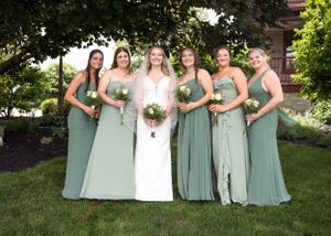 A bridal party poses outdoors wearing sage green bridesmaid dresses against a landscaped garden backdrop.