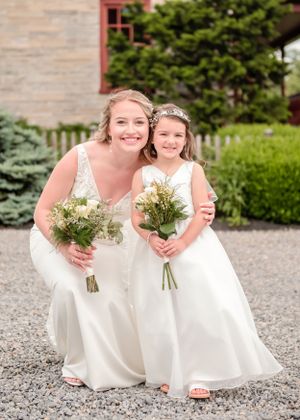Two figures in white wedding dresses hold small bouquets of wildflowers while posing together on gravel path by garden.