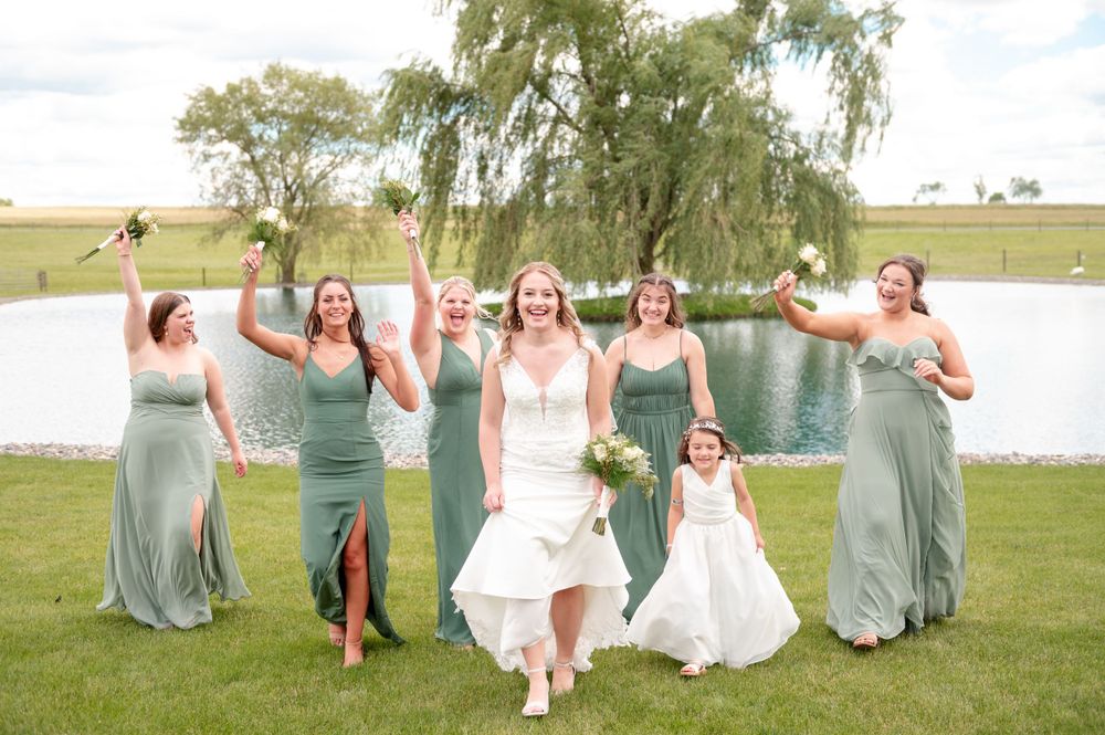 Celebratory group in sage green dresses and white gown pose joyfully by a lake under a weeping willow tree.