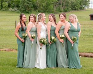 A group of bridesmaids in sage green dresses stand together holding bouquets in an outdoor field setting.
