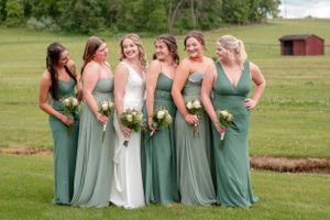 Wedding party wearing elegant sage green bridesmaid dresses pose together on grassy field with rustic barn in background.