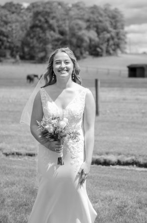 Black and white portrait of bride holding bouquet in countryside meadow with barn and trees in background.