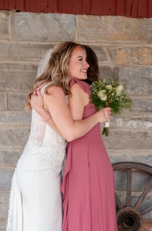 Two people share an emotional embrace at a wedding, with one in a white gown and another in a dusty rose dress holding flowers.
