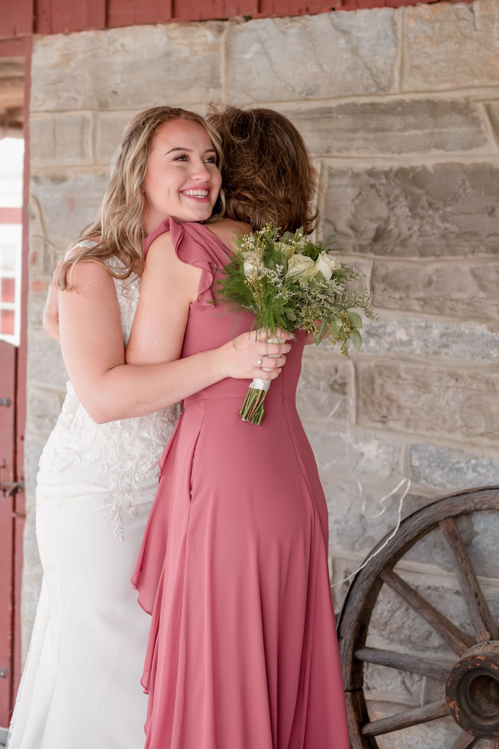 Two women share an emotional embrace at a wedding, with one in white lace and one in a dusty rose dress holding a bouquet.