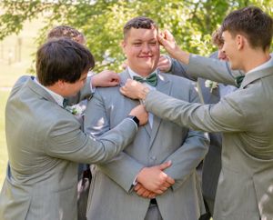 Groomsmen in matching light gray suits playfully adjust the groom's bow tie and tuxedo outdoors.