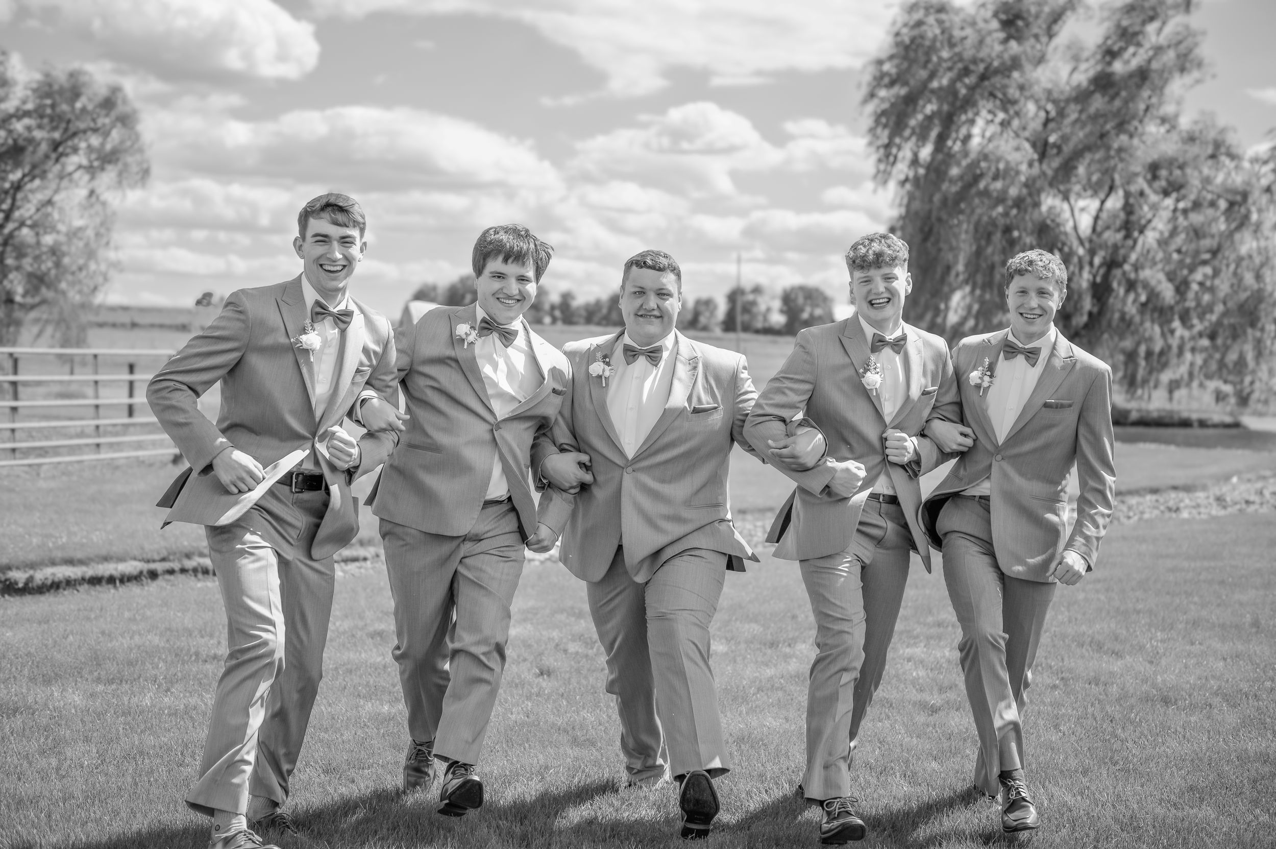 Wedding party in matching gray suits pose playfully while kicking their legs up on a grassy lawn in black and white.