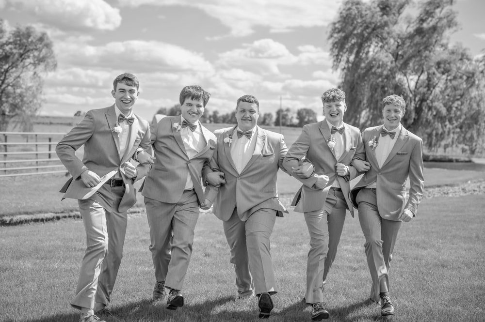 Wedding party in matching gray suits pose playfully while kicking their legs up on a grassy lawn in black and white.