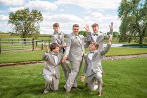 A playful group wearing light gray suits poses and jumps on a manicured lawn with a fence in the background.