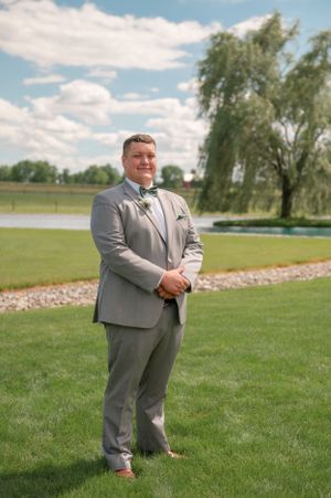 Groom in light grey suit and bowtie standing outdoors on sunny day with blue sky.