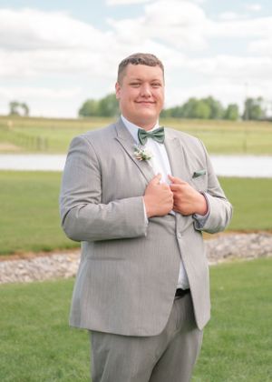 A groom in a light grey suit and bowtie smiles while adjusting his jacket in an outdoor setting.