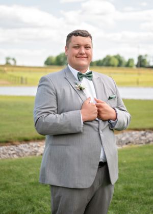 A person in a light gray suit with a green bow tie stands outdoors by a lake on a grassy lawn.