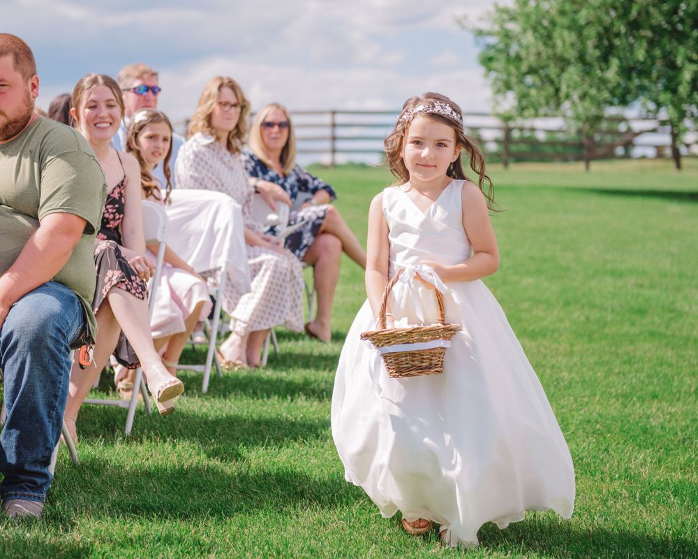 A flower girl in a white dress walks down a grassy aisle at an outdoor wedding ceremony, carrying a small basket.