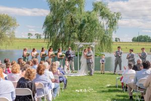 Outdoor wedding ceremony under a willow tree beside a lake with guests seated in white chairs.