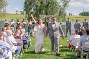 A joyful wedding celebration takes place outdoors under a weeping willow tree with guests seated on white chairs.