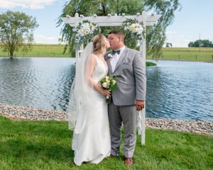 Newlyweds share a romantic moment under a white floral arch with a scenic lake backdrop.