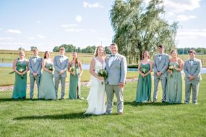 Wedding party in mint green and gray attire poses together on a sunny day at an outdoor venue with grassy grounds.
