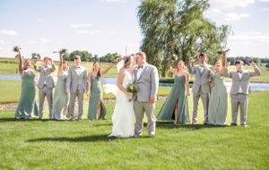 Wedding party in sage green and gray attire poses on lawn with lake and willow tree backdrop.