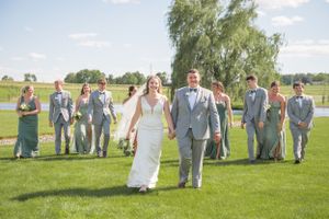 Wedding party walking outdoors on manicured lawn with bridesmaids in blue dresses and groomsmen in gray suits.