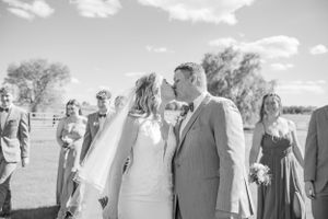 A wedding party gathers outdoors on a sunny day with white clouds in the sky and trees in the background.