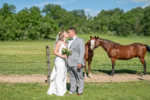 Wedding couple shares romantic moment in field with brown horse watching in background.