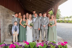Wedding party in sage green and gray attire poses together under rustic barn overhang with pink flowers.
