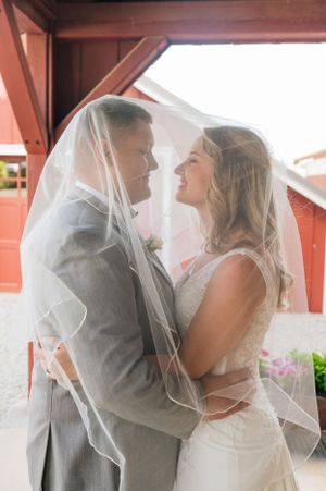 A romantic moment between newlyweds sharing an intimate embrace under a wedding veil at a rustic red barn venue.