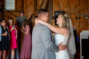 A couple enjoys their first dance as newlyweds inside a rustic wooden barn while wedding guests look on.