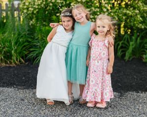 Three young girls in white and pastel dresses smile together against landscaped garden background.