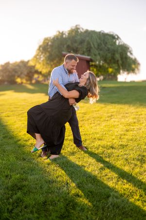 Romantic couple dips and embraces during golden hour sunset on grassy hillside.