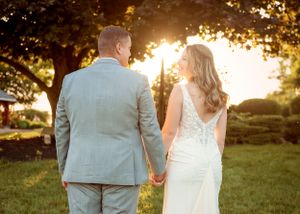 A couple in wedding attire holds hands while walking together at sunset in a scenic garden setting.