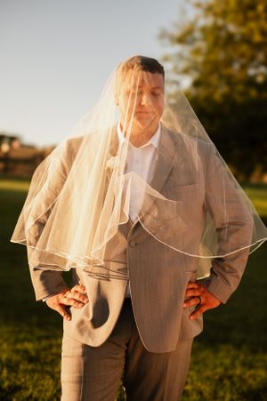 Groom wearing a light suit poses playfully with bride's veil in golden sunset light.