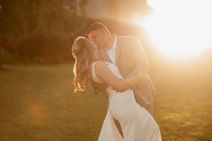 A couple in wedding attire embrace during golden hour sunset in a romantic outdoor setting.