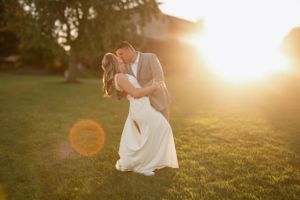 A romantic wedding photo in a grassy field at golden hour with sun flare and warm backlighting.