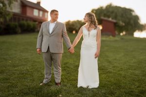 Bride in white dress and groom in grey suit holding hands in garden at sunset.