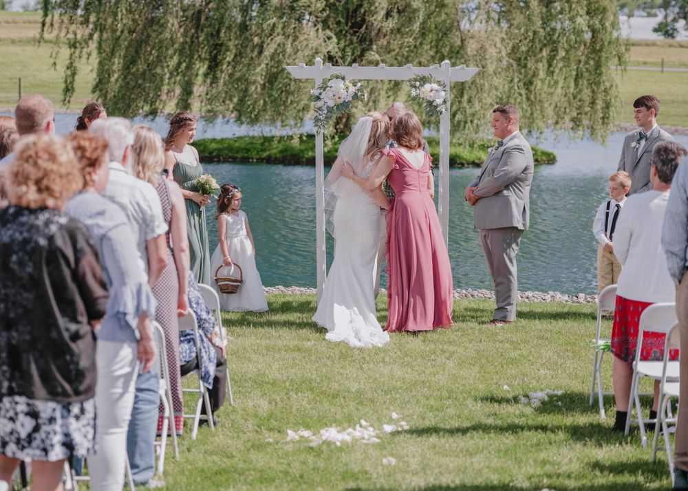 An outdoor wedding ceremony takes place under a white arch by a lake with a weeping willow tree in the background.