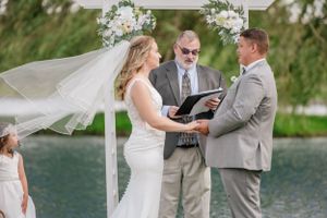 Outdoor waterfront wedding ceremony with flowing white veil and floral arch overlooking a serene lake.
