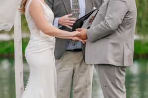 Close-up of couple holding hands during outdoor wedding ceremony with officiant in grey suit reading from book.
