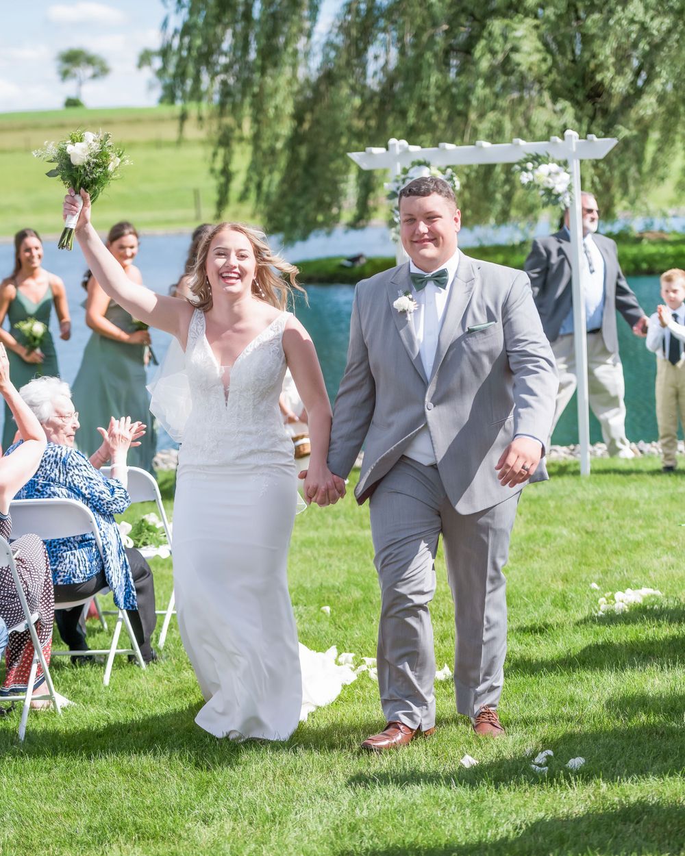 Joyful couple celebrates during their wedding ceremony under a floral arch beside a lake on a sunny day.