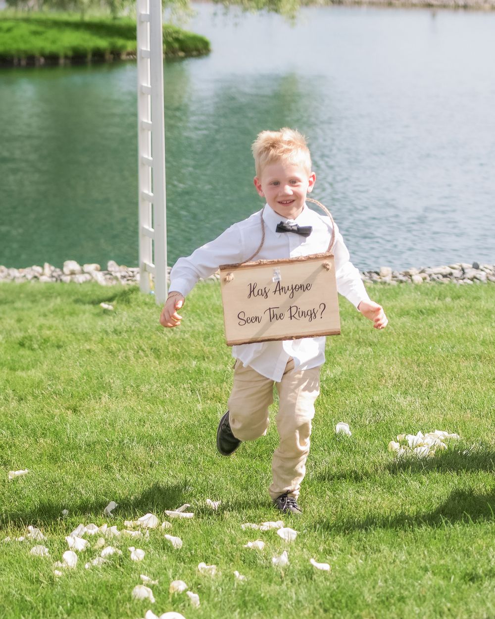 A joyful child in a bowtie runs across green grass by a lake carrying a rustic wooden sign.