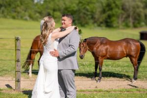 Newlyweds share romantic moment in field with brown horse grazing in background.