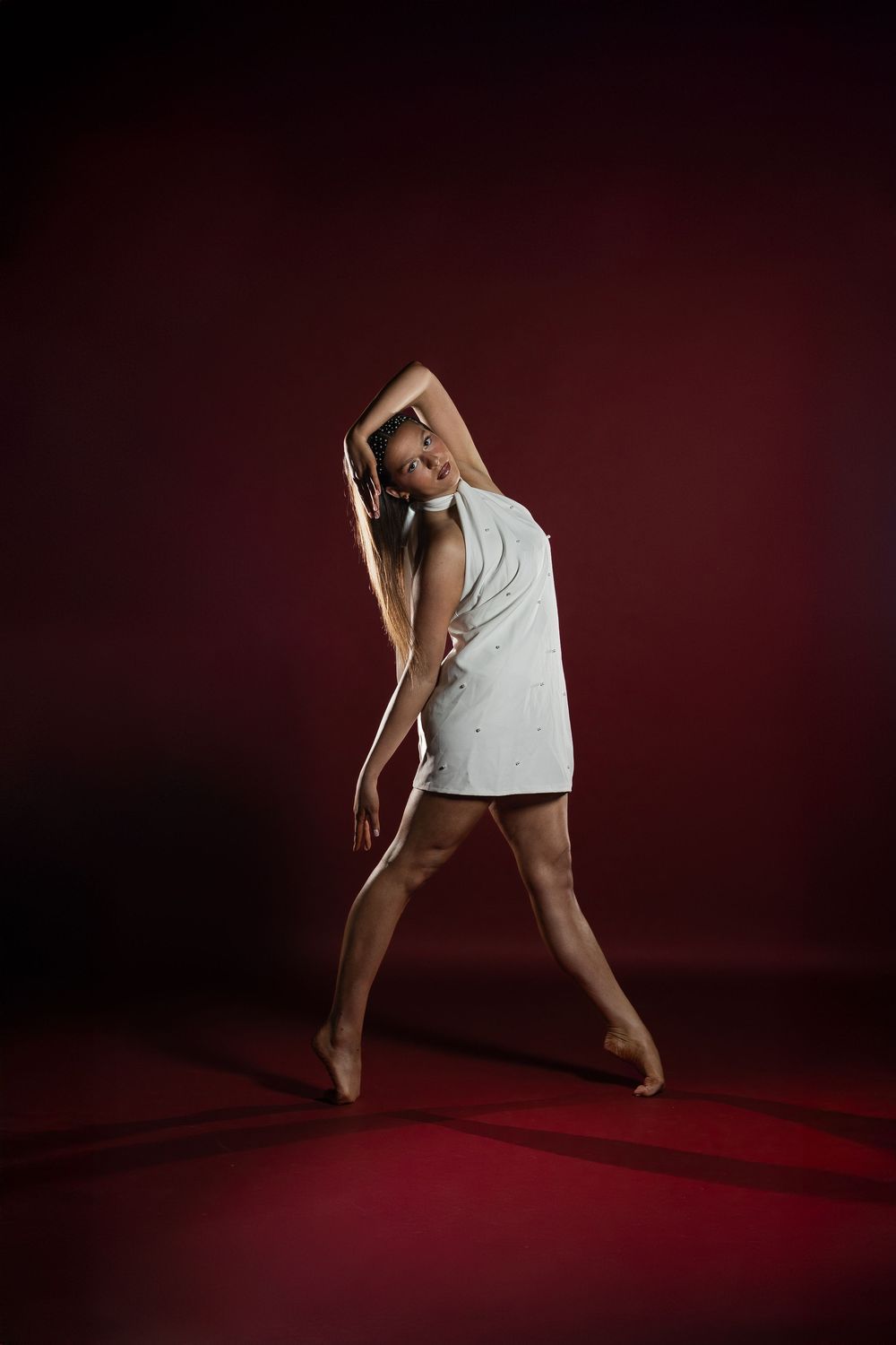 Contemporary dancer in white top performs expressive floor movement sequence against dark red backdrop.