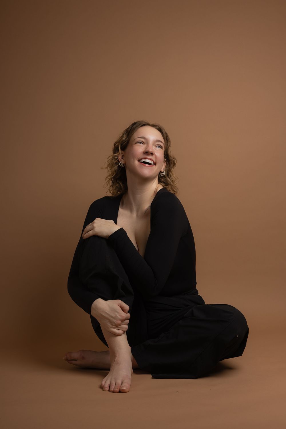 A laughing woman in a black jumpsuit sits cross-legged on the floor against a warm beige background.