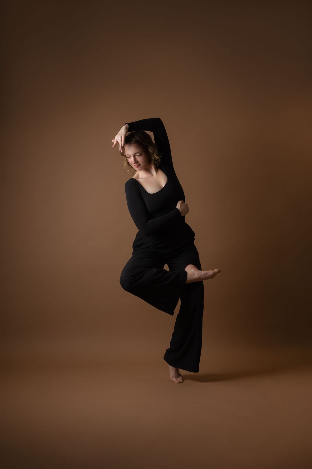 A barefoot dancer in black attire poses gracefully on one leg against a warm brown studio background.