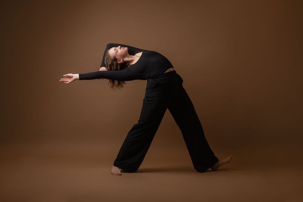 A dancer in black attire performs an expressive contemporary dance pose against a warm brown studio background.