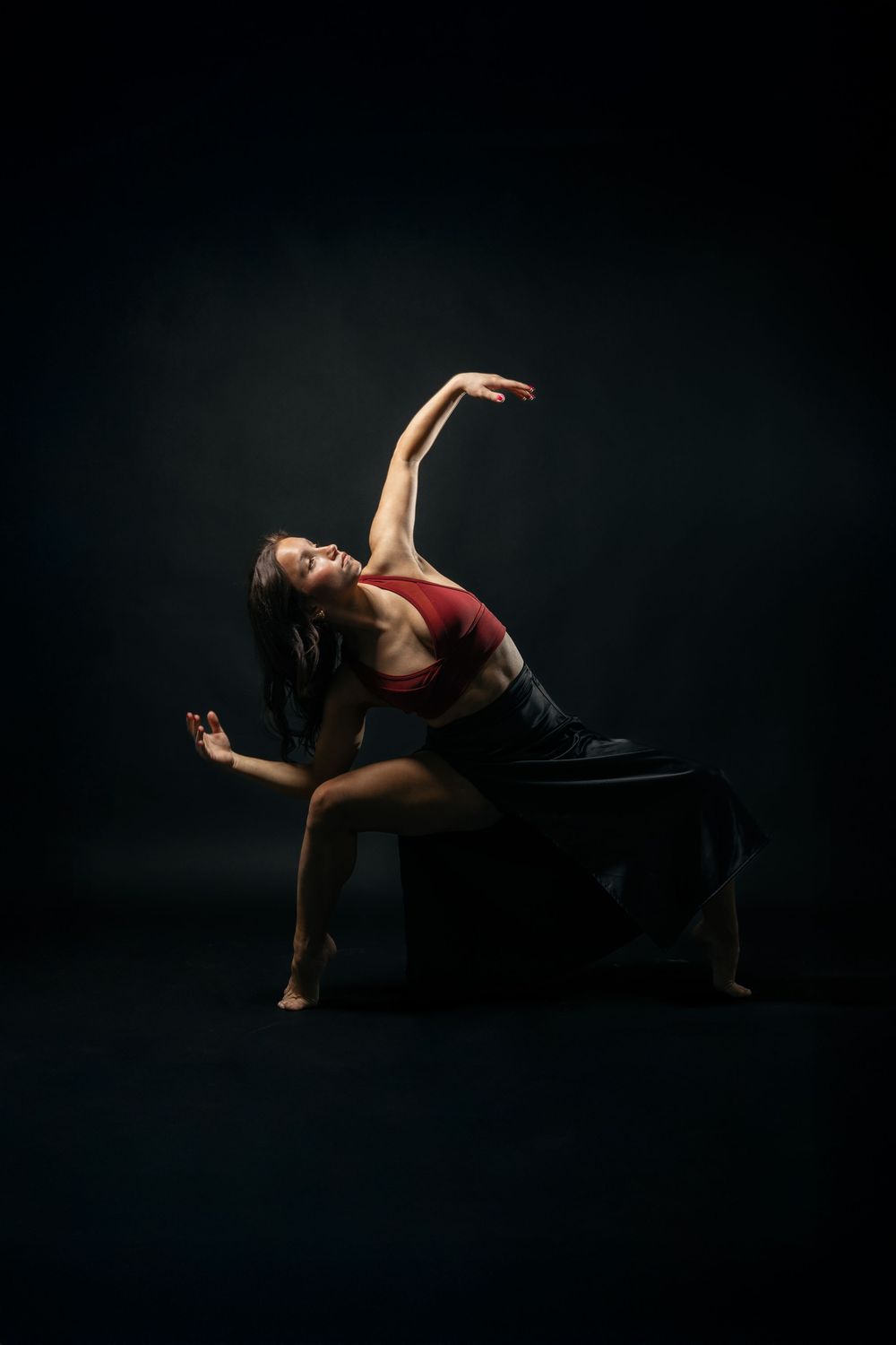 Dancer in red top performs graceful pose on dark stage, arching backwards in dramatic lighting against black background.