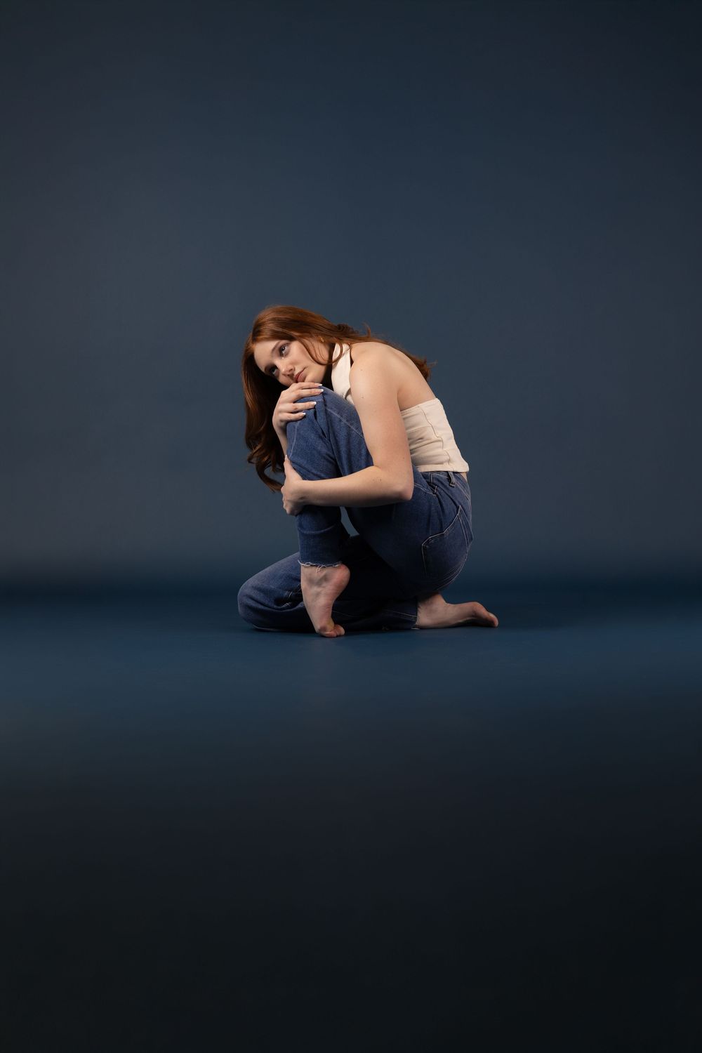 A young woman with long red hair sits on the floor hugging her knees, wearing jeans and a white top on a dark blue background.