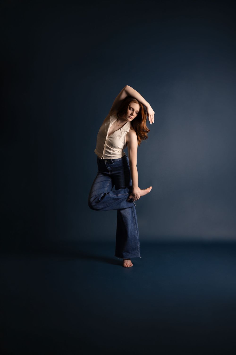 A barefoot woman in cream tank top and blue wide-leg pants poses gracefully in a dark studio setting.