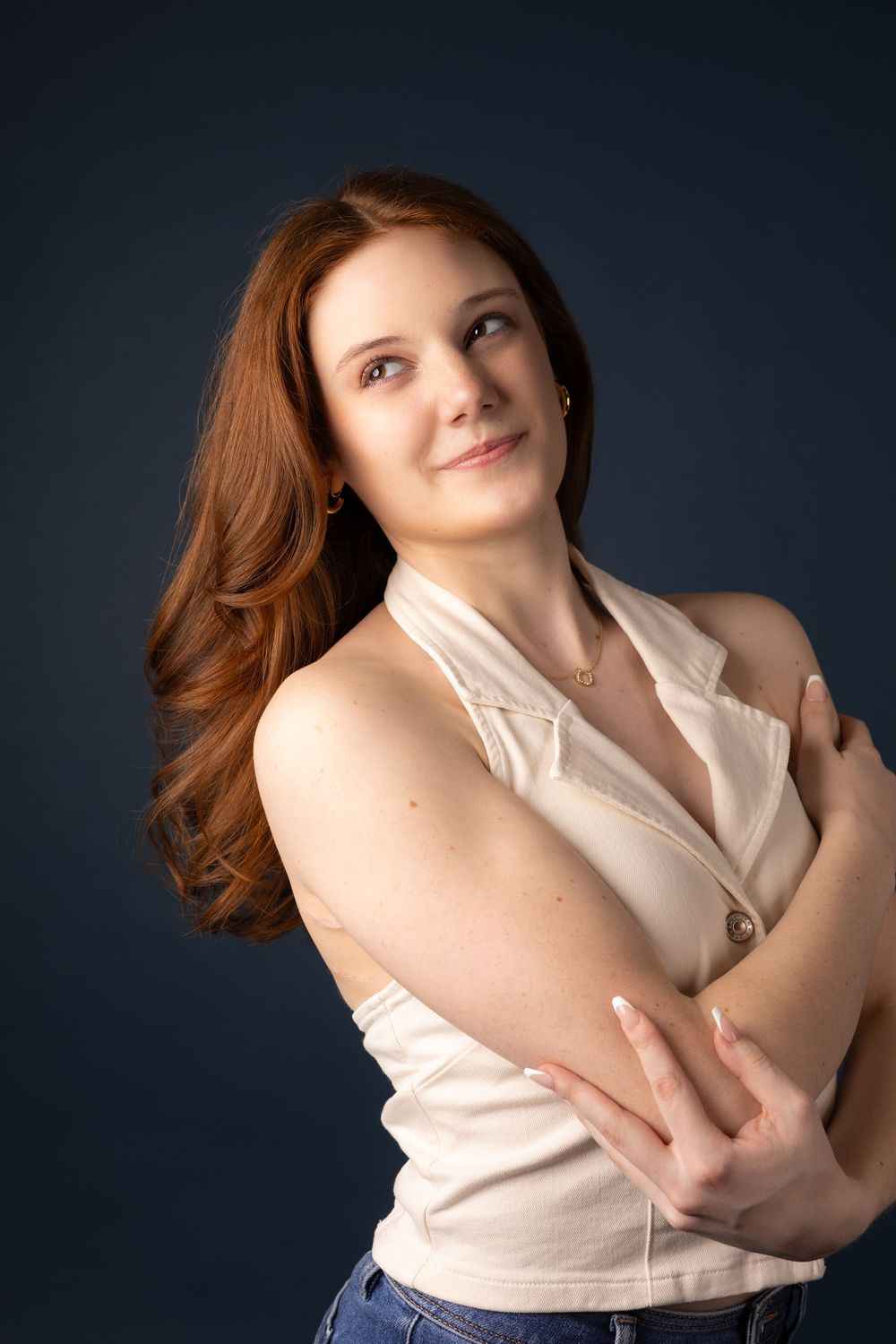Young woman with long red hair in white sleeveless top and jeans, arms crossed, smiling against dark blue background.