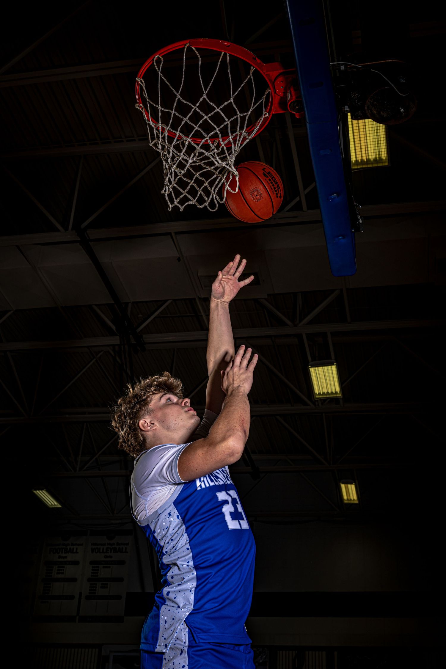 Basketball player in blue jersey leaps toward hoop for layup under dramatic lighting.